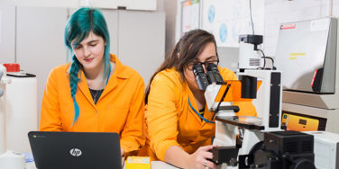 two science students look through microscope and at a laptop